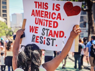 The cover of GCIR's report, Immigration Legal Services in California: A Time for Bold Action, featuring a woman holding a sign reading, "This is America. United We Stand. Resist."