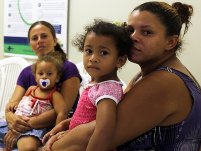 Two women with young girls on their laps, one with a pacifier, awaiting doctor's appointments in an office. Posted to accompany GCIR's brief and funding recommendations, Protecting Families and Advancing Belonging: How Philanthropy Can Answer Threats to the Well-Being of Immigrants.